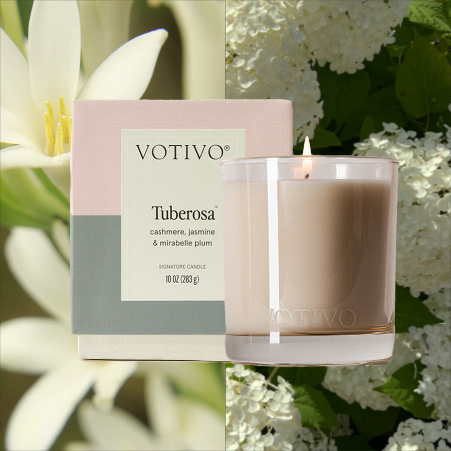 A Votivo Tuberosa 10 oz. Signature Candle and its box are side by side; behind them, white tuberose flowers fill one half of the background, while white jasmine blooms with green leaves cover the other half.