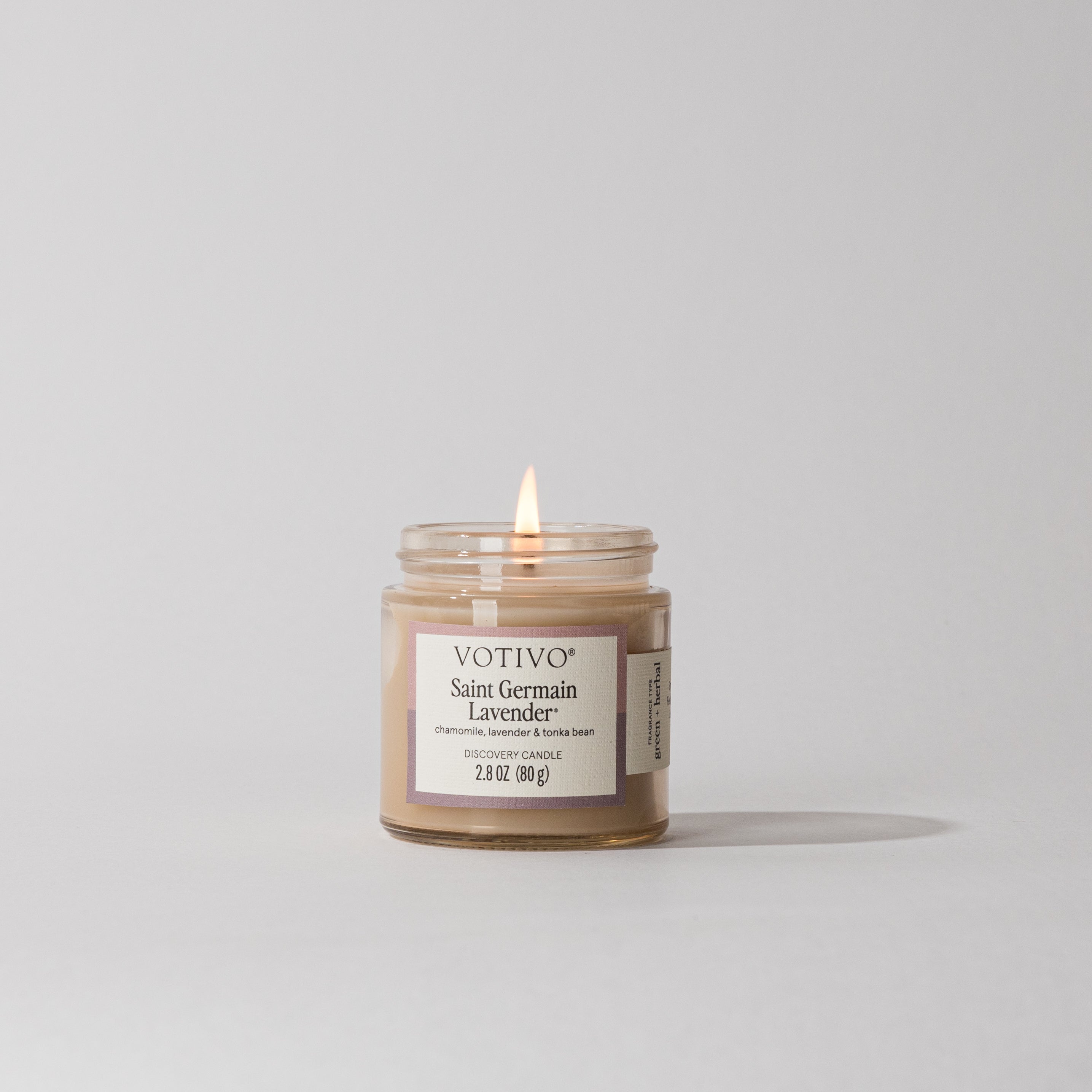 A Votivo Saint Germain Lavender Discovery Candle, featuring creamy beige soy blend wax and a gentle flame, is set against a plain light gray background.