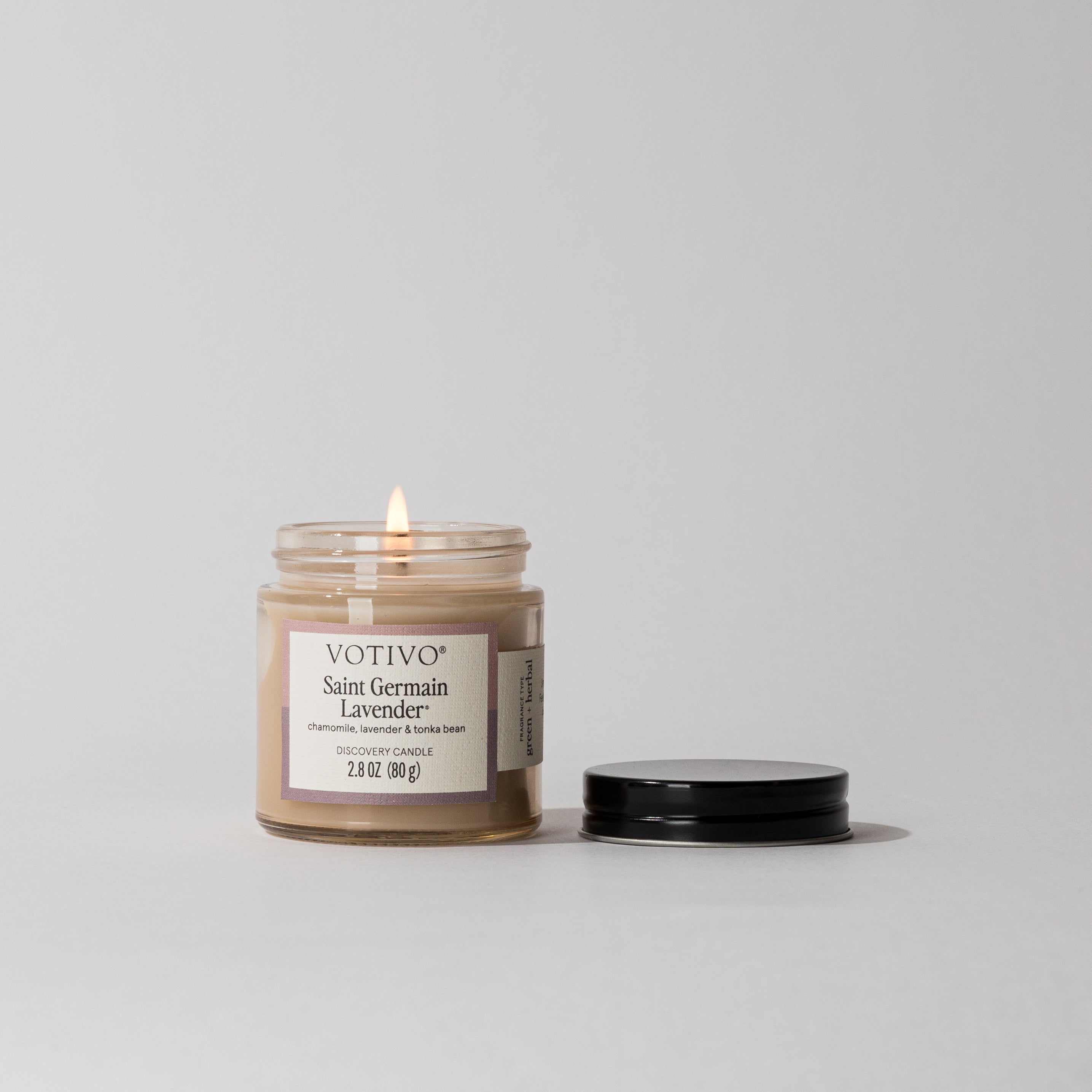 A lit Saint Germain Lavender Discovery Candle by Votivo, made with soy blend wax, sits in a clear glass jar on a white surface with its black lid beside it.