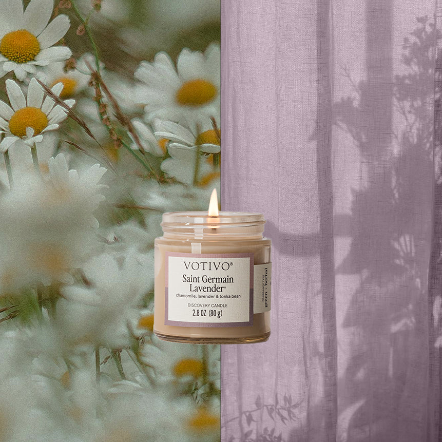 A glowing Votivo Saint Germain Lavender Discovery Candle sits at the center, framed by blooming daisies on the left and sheer lavender curtains with soft shadows on the right.