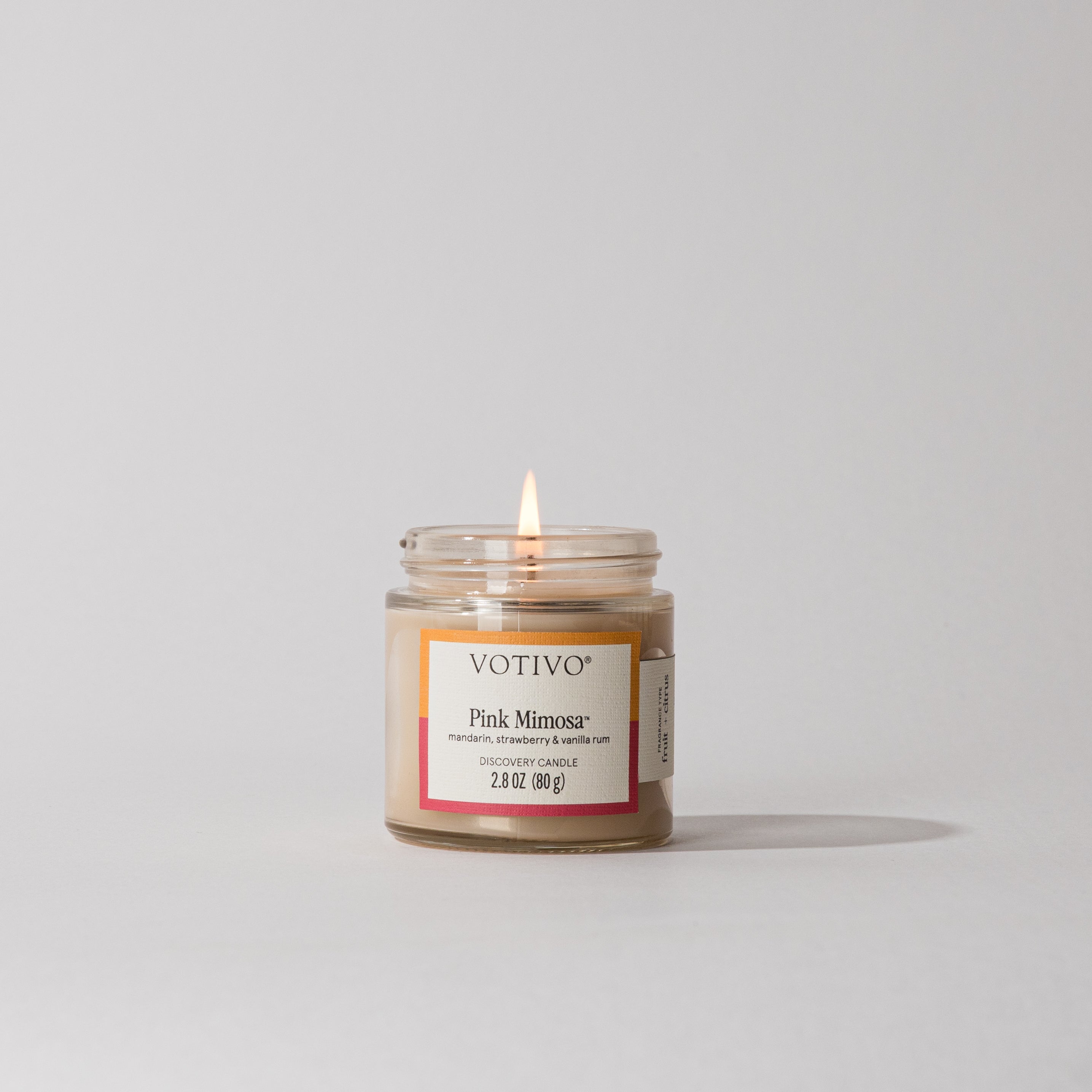 A Votivo Pink Mimosa Discovery Candle glows in a clear glass jar on a white surface. The label mentions mandarin, strawberry, and vanilla rum scents, and the candle is made from a soy blend wax.