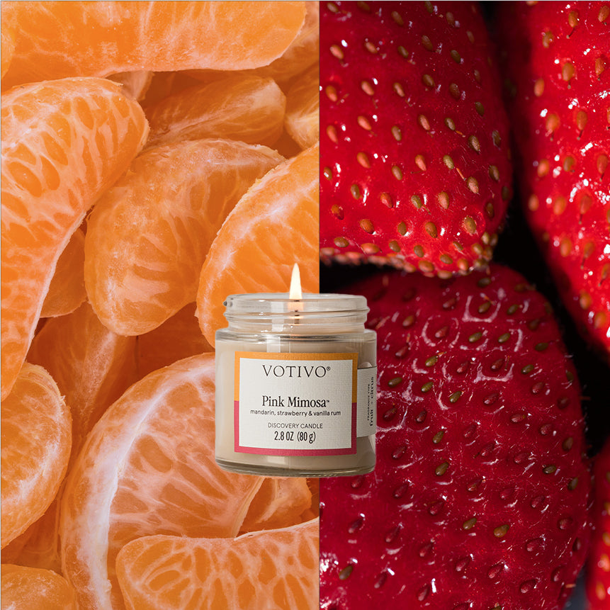A Votivo Pink Mimosa Discovery Candle is lit, set before a split background with tangerine segments on the left and strawberries on the right.