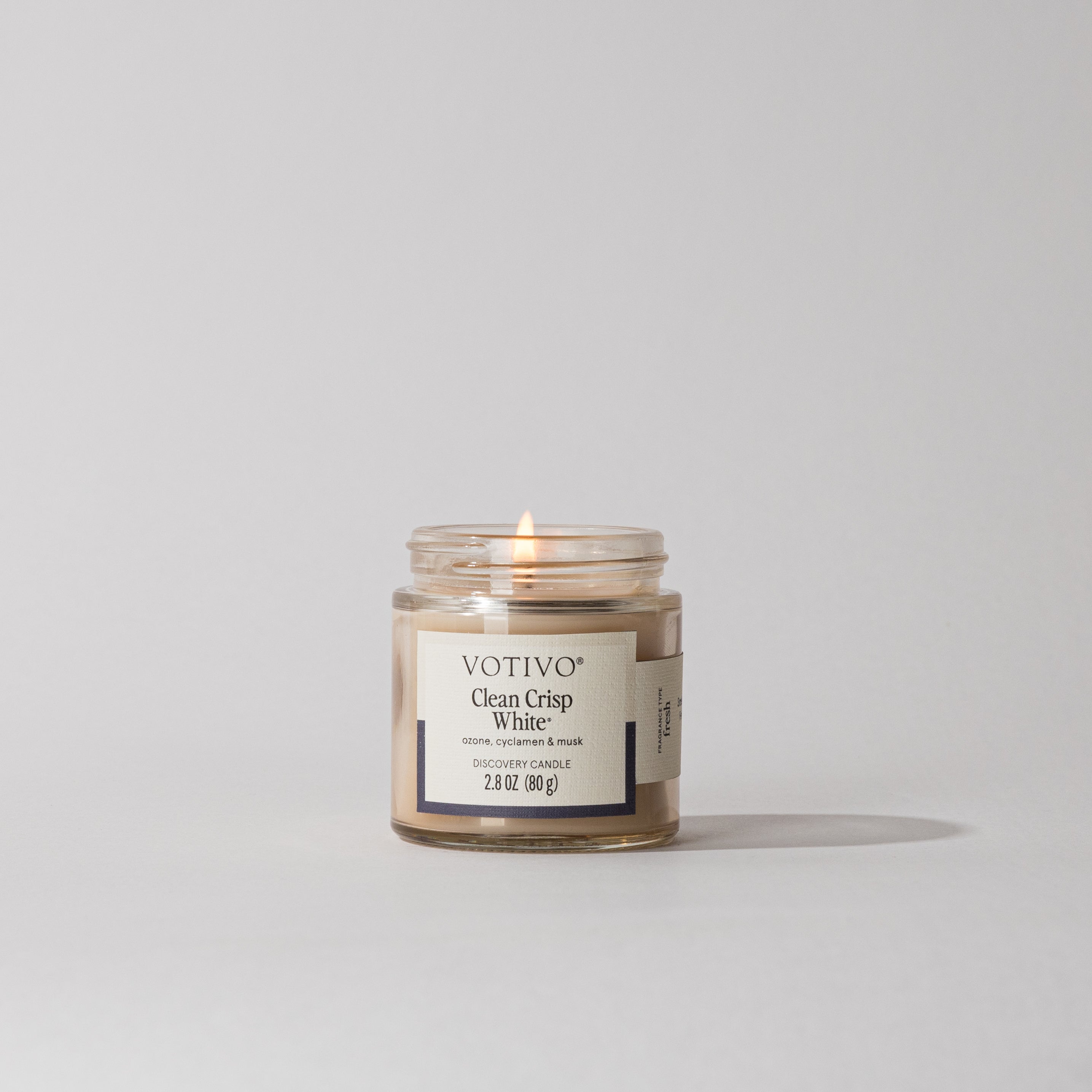 A Votivo Clean Crisp White Discovery Candle with soy blend wax burns gently in a small glass jar against a plain light gray background.