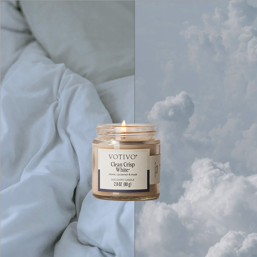 A lit Votivo Clean Crisp White Discovery Candle is placed between a soft, rumpled white bed on the left and a sky filled with fluffy clouds on the right.