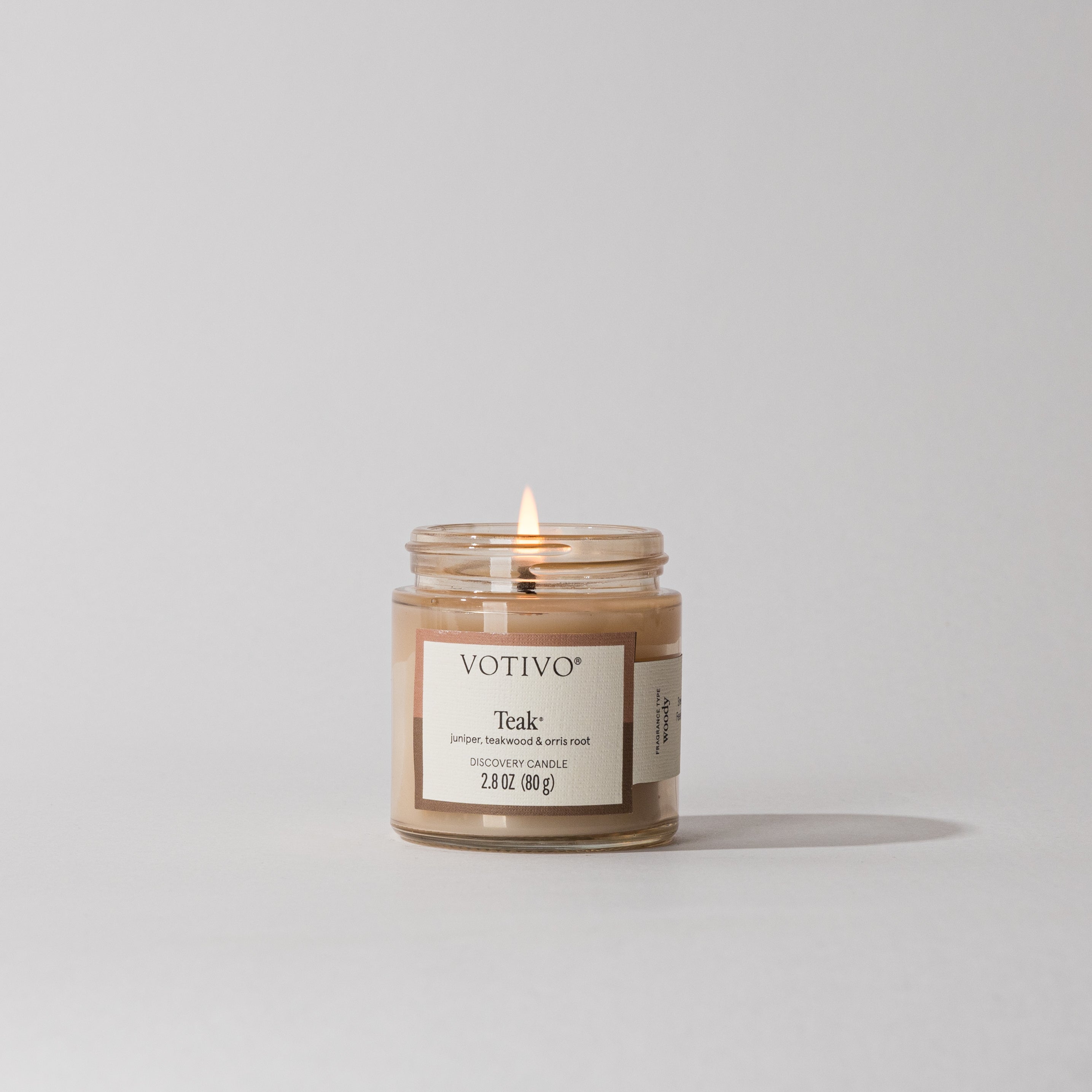 A small Votivo Teak Discovery Candle glows with a gentle flame, filling the area with a warm, woody scent. The candle sits against a plain light gray background.