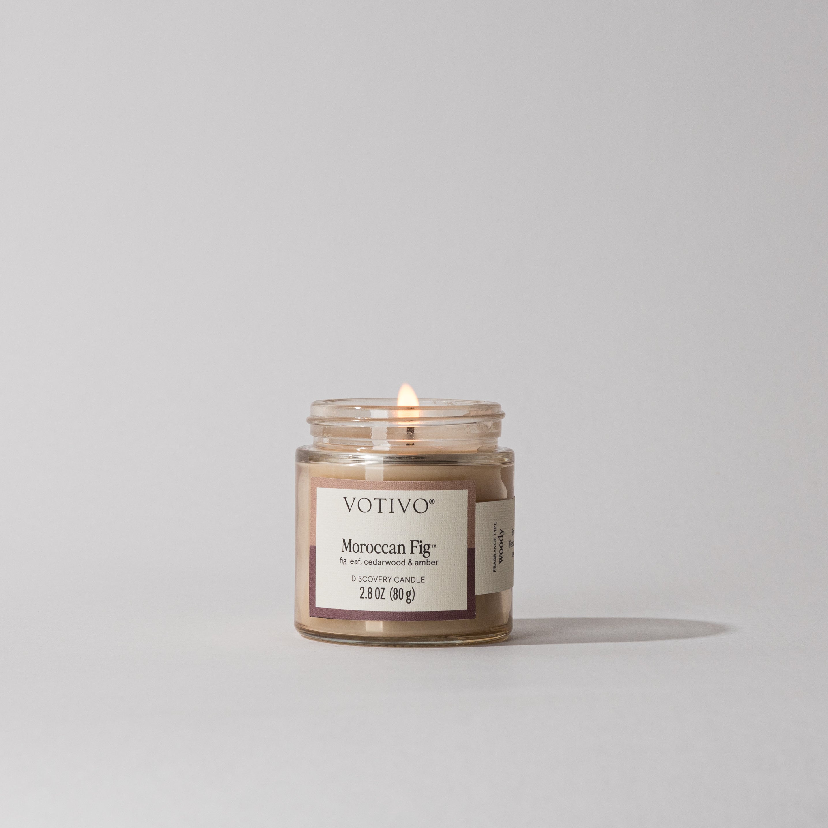 The Moroccan Fig Discovery Candle by Votivo features beige soy blend wax and a minimalist glass jar, burning with a single flame against a light gray background.
