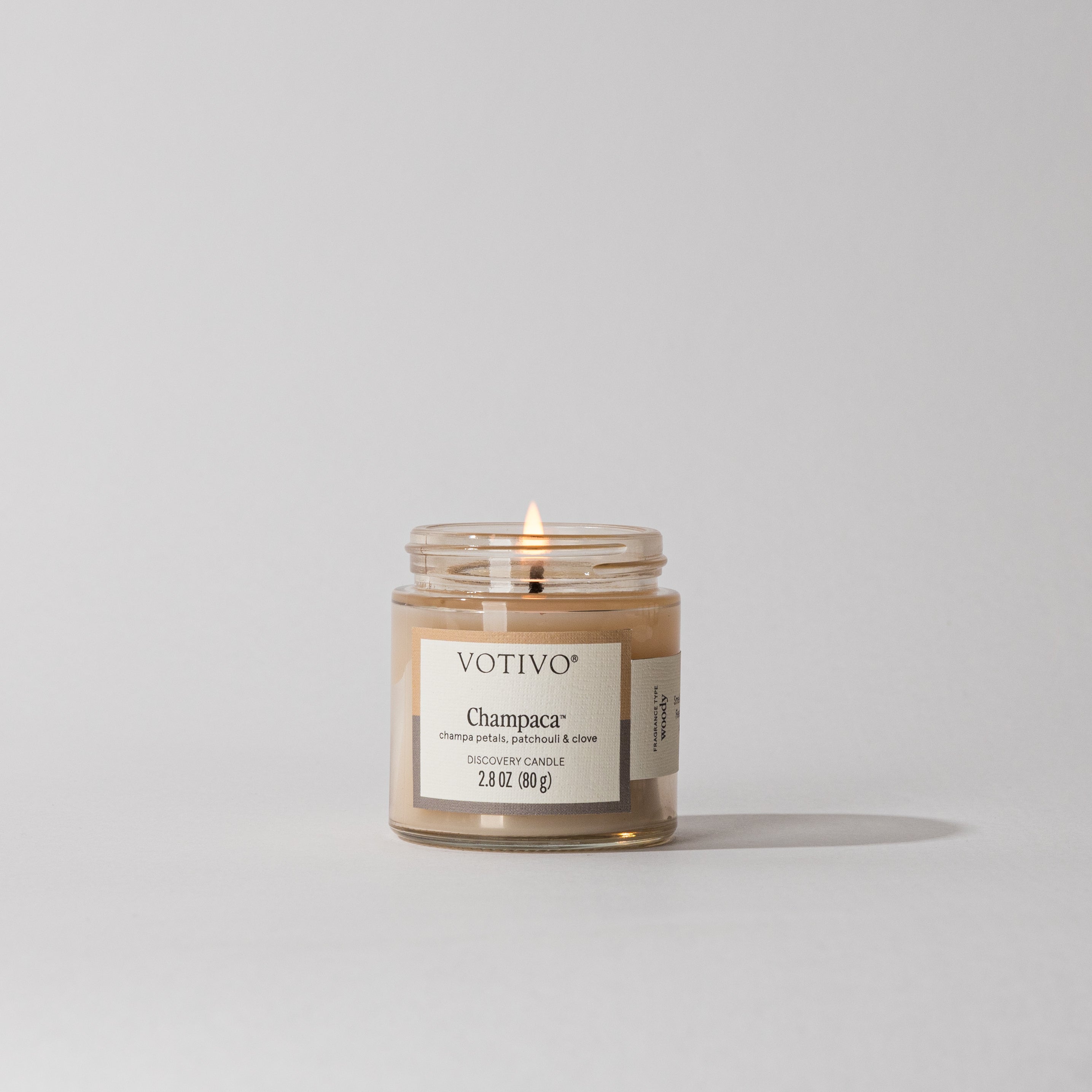 A Votivo Champaca Discovery Candle with beige soy-blend wax burns with a single flame against a plain light gray background, featuring a minimalist design and filling the air with its subtle woody scent.
