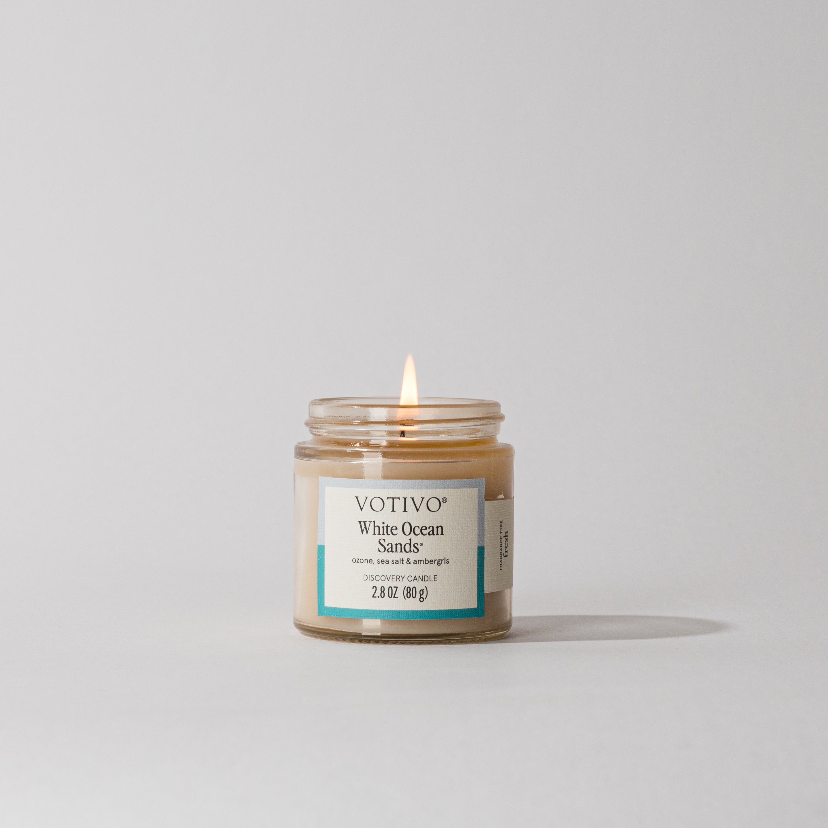 A lit Votivo White Ocean Sands Discovery Candle in a glass jar rests on a plain light gray background, crafted from premium soy blend wax.