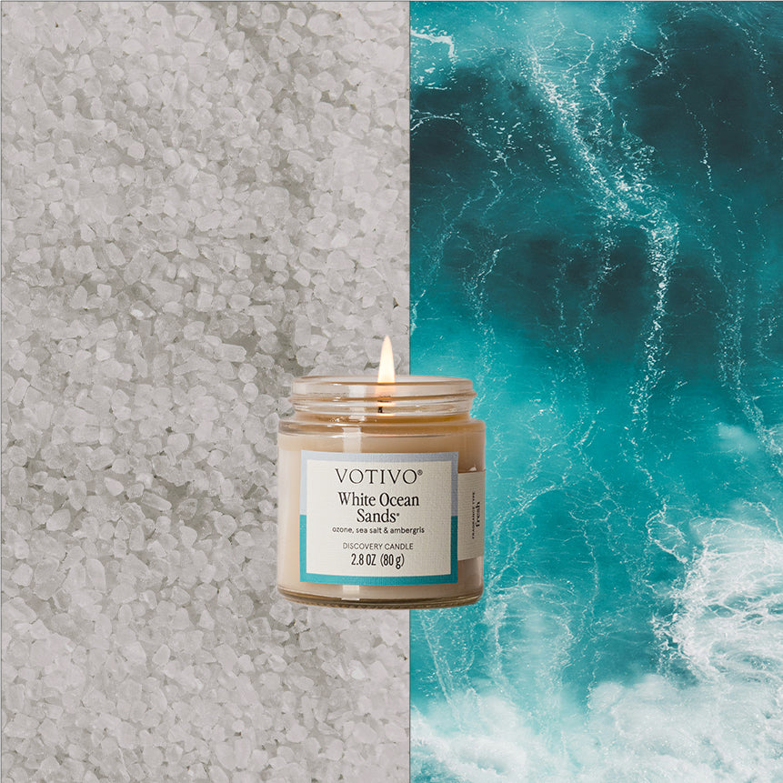 A lit Votivo White Ocean Sands Discovery Candle sits against a split backdrop: coarse grey salt on the left and vivid blue ocean waves on the right.