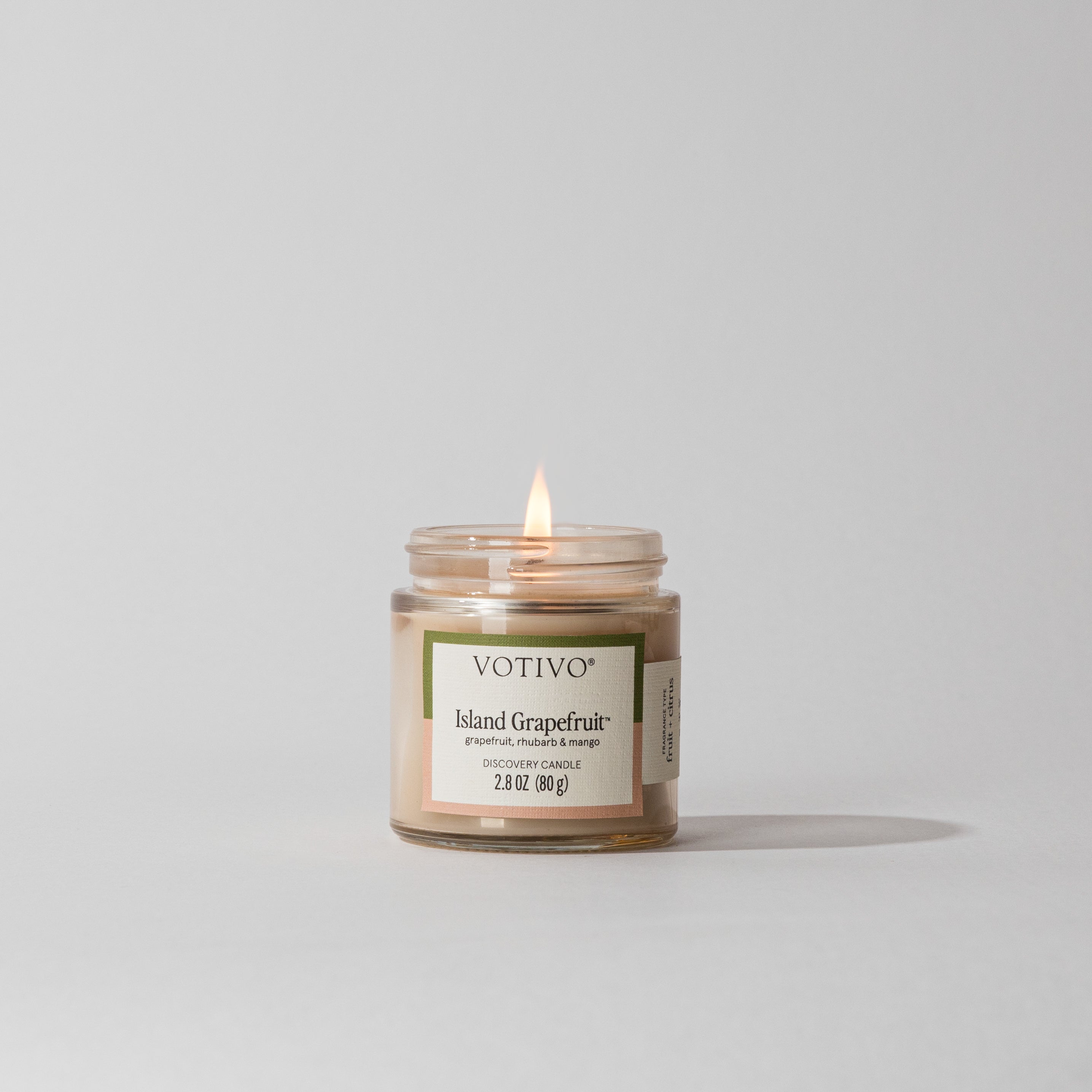 A Votivo Island Grapefruit Discovery Candle glows in a clear glass jar against a white background. Made with soy blend wax, its label features notes of grapefruit, rhubarb, and tangerine.