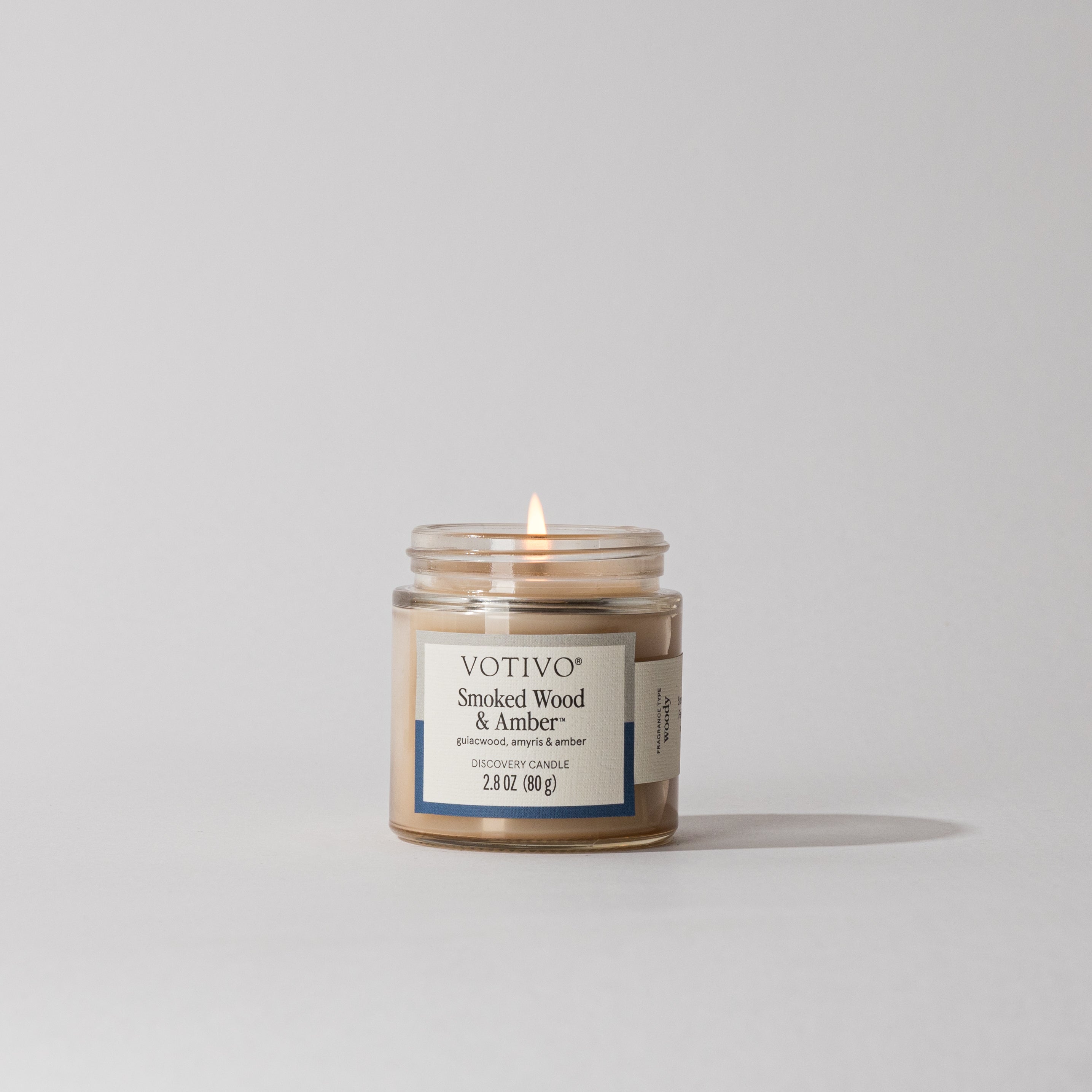 A lit Votivo Smoked Wood & Amber Discovery Candle rests on a plain white surface, its soy blend wax casting a gentle shadow from the small glass jar.