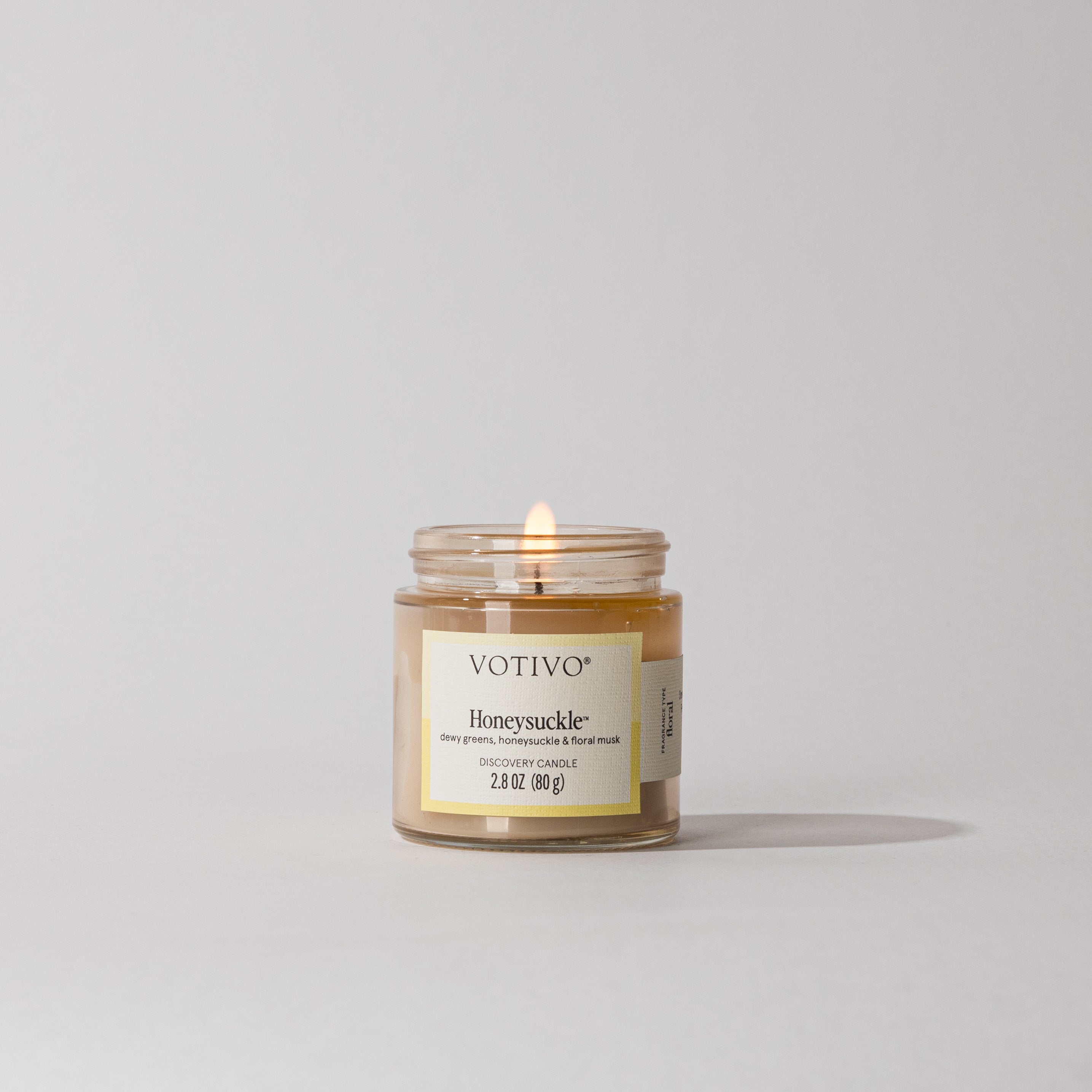 A lit Votivo Honeysuckle Discovery Candle with yellow wax and label sits in a clear glass jar on a light gray surface, its delicate floral fragrance gently filling the room.