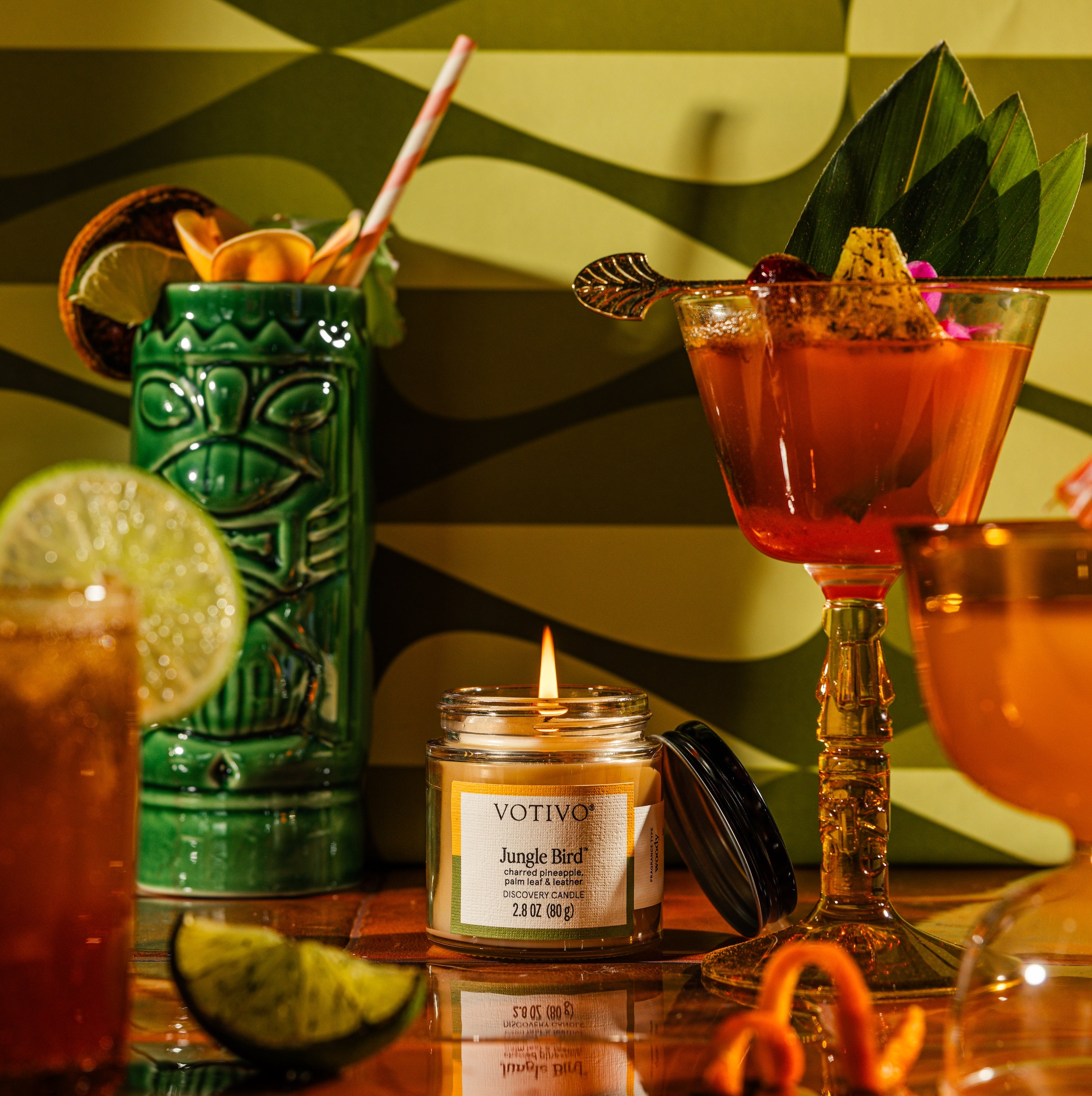 A lit Votivo Jungle Bird Discovery Candle sits amid bright tropical cocktails, garnishes, and citrus slices on a green wavy background, creating a festive summer vibe with Votivos signature long-lasting fragrance.