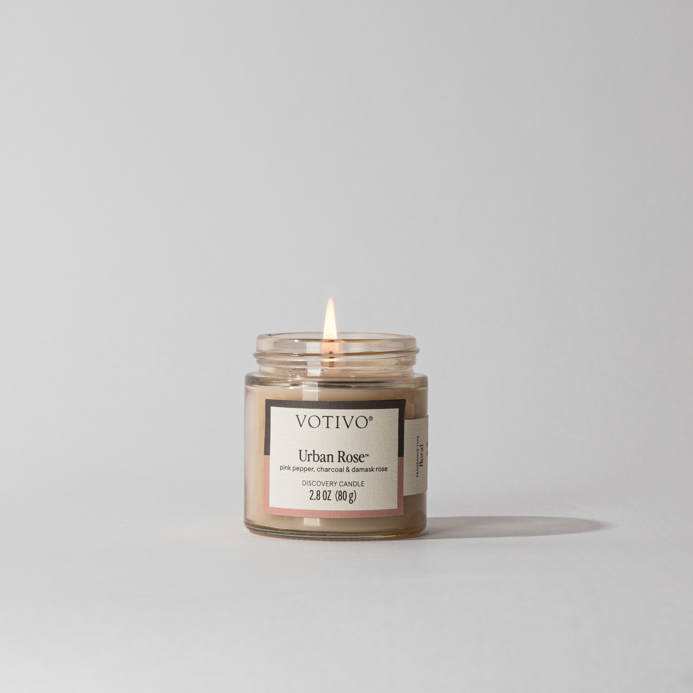A Votivo Urban Rose Discovery Candle, made with premium soy blend wax, sits in a clear glass jar on a plain light surface with a neutral background. Its label, featuring blush pink accents and text, is clearly visible.