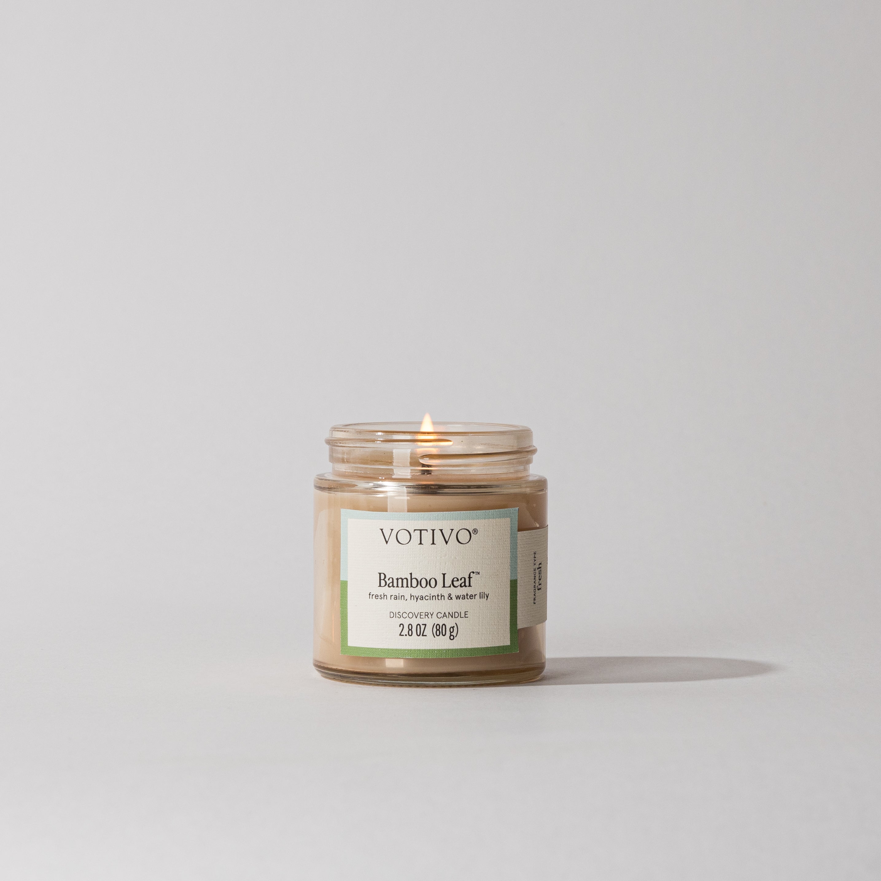 A Votivo Bamboo Leaf Discovery Candle, made with clean-burning soy blend wax, glows in a clear glass jar on a light gray surface, casting a soft shadow against a plain, light-colored background.