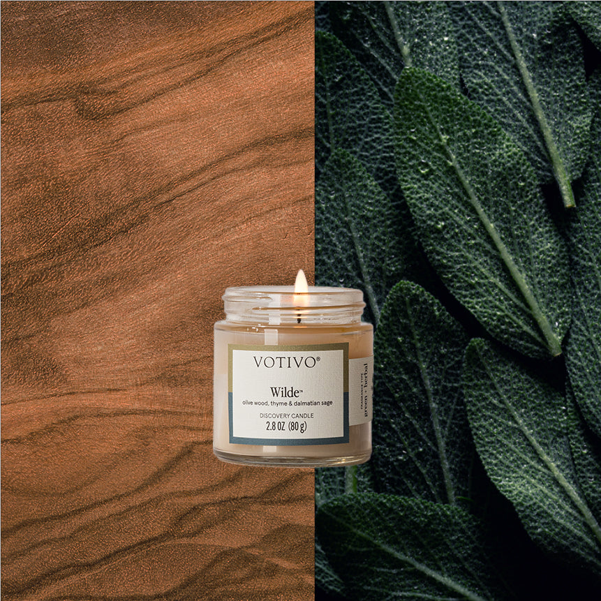 A Votivo Wilde Discovery Candle is centered, flanked by a wood grain backdrop on the left and large, textured green leaves to the right.