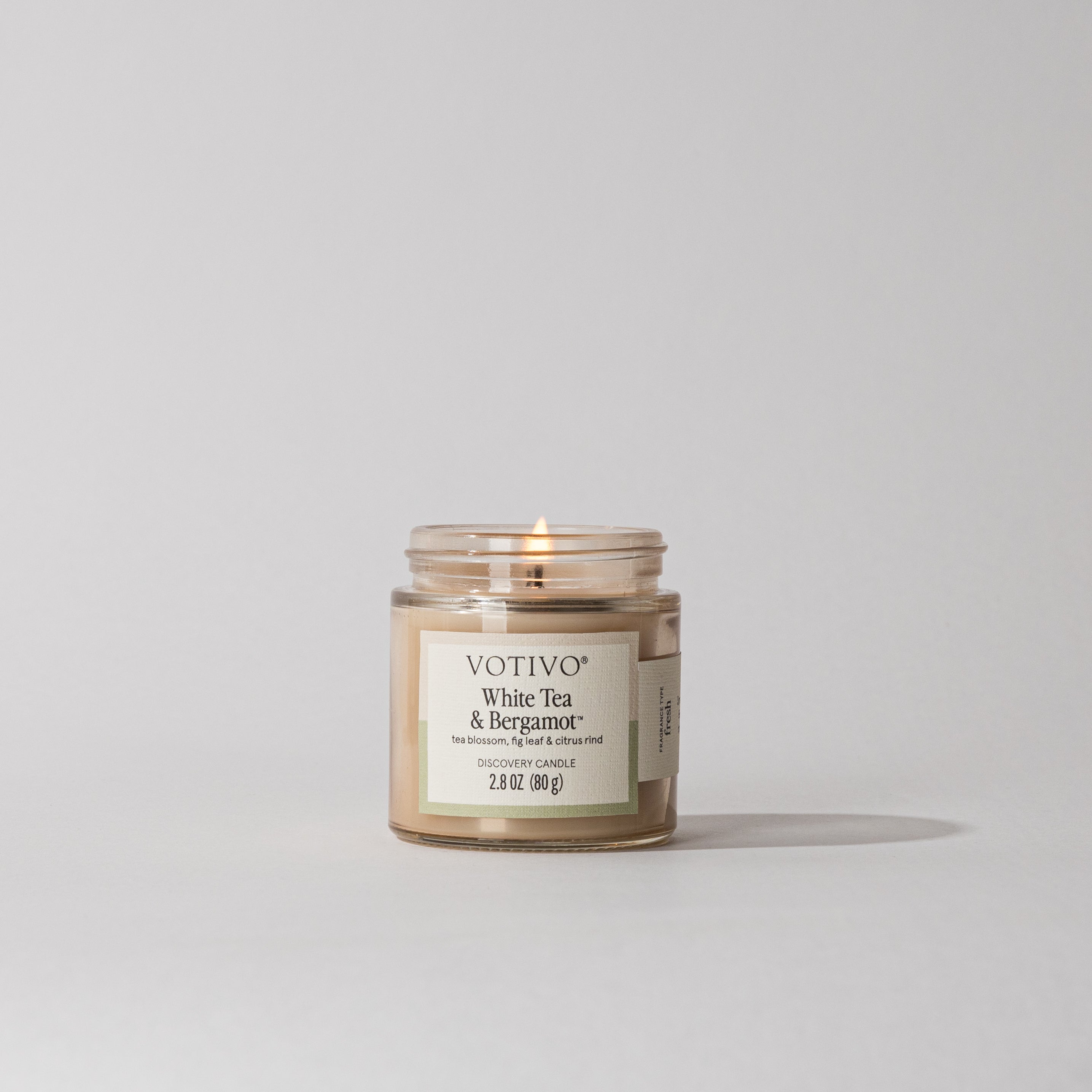 A lit Votivo White Tea & Bergamot Discovery Candle sits in a small glass jar. The label displays scent details, weight (2.8 oz / 80 g), and premium soy blend wax. The background is plain light gray.