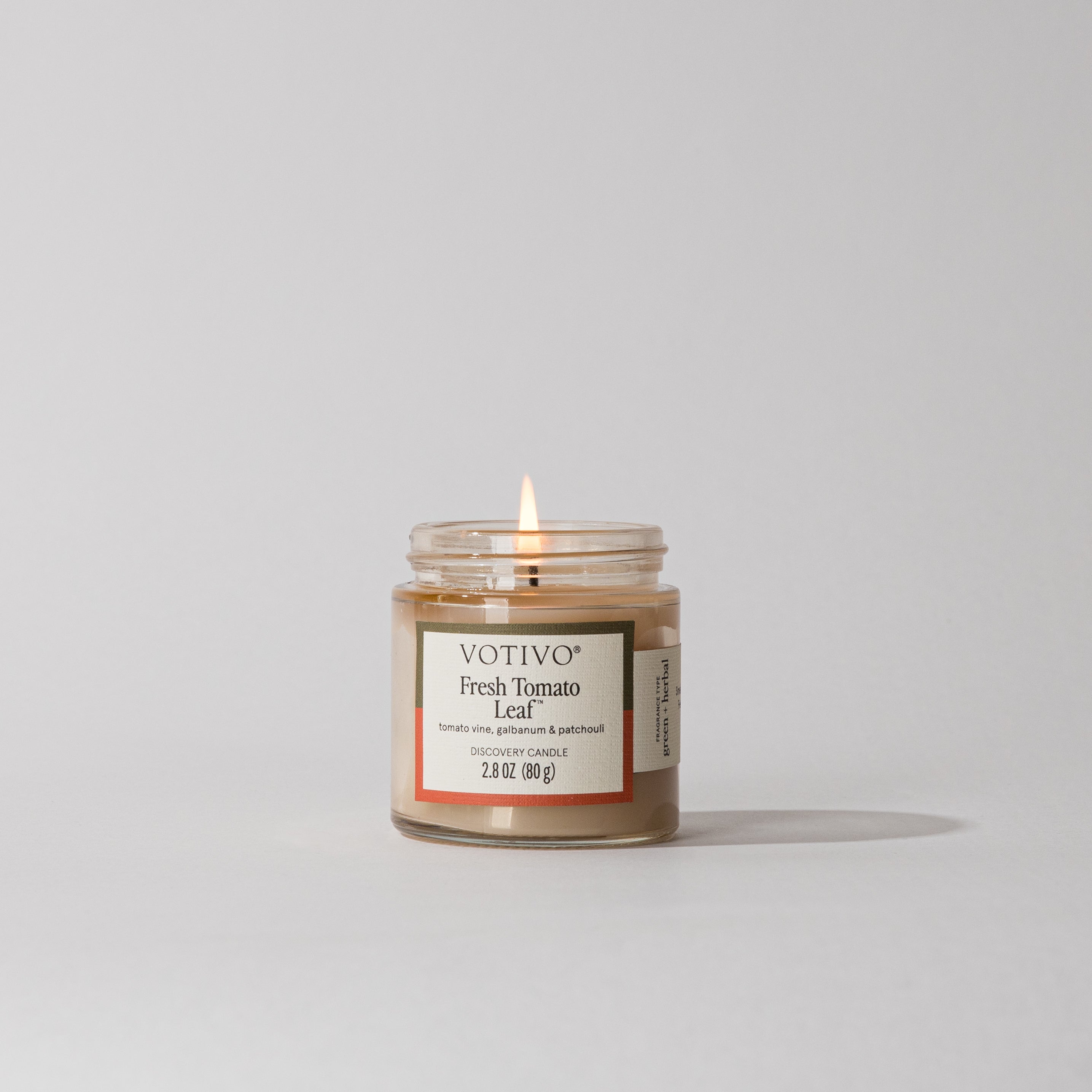 The Votivo Fresh Tomato Leaf Discovery Candle, a small glass jar candle, emits a steady flame and fills the air with fresh tomato leaf, galbanum, and patchouli scents from its soy blend formula against a plain light background.