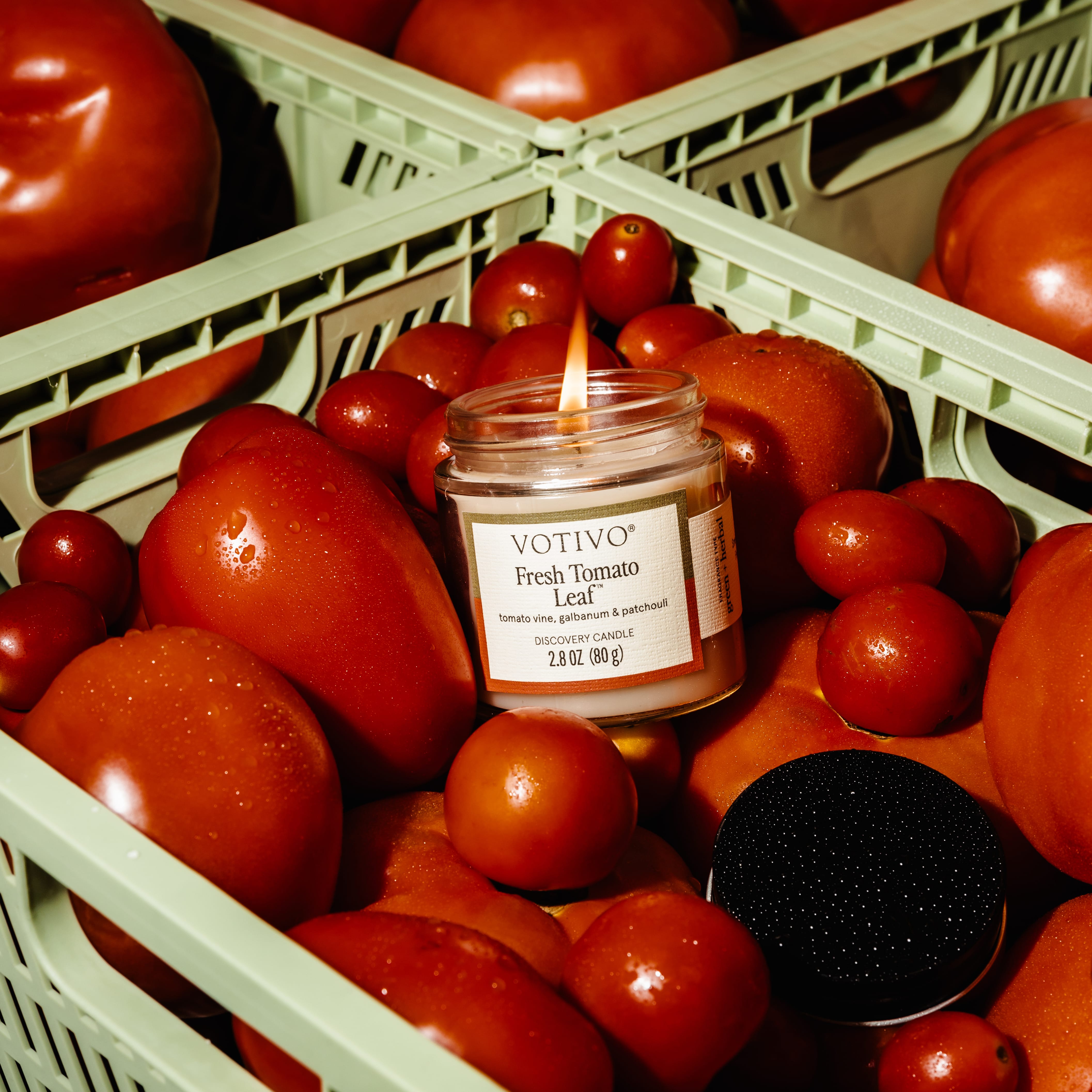 A lit Votivo Fresh Tomato Leaf Discovery Candle in a glass jar rests among ripe tomatoes in a green crate. The soy blend candles black lid lies nearby, while its fresh tomato leaf fragrance fills the air.