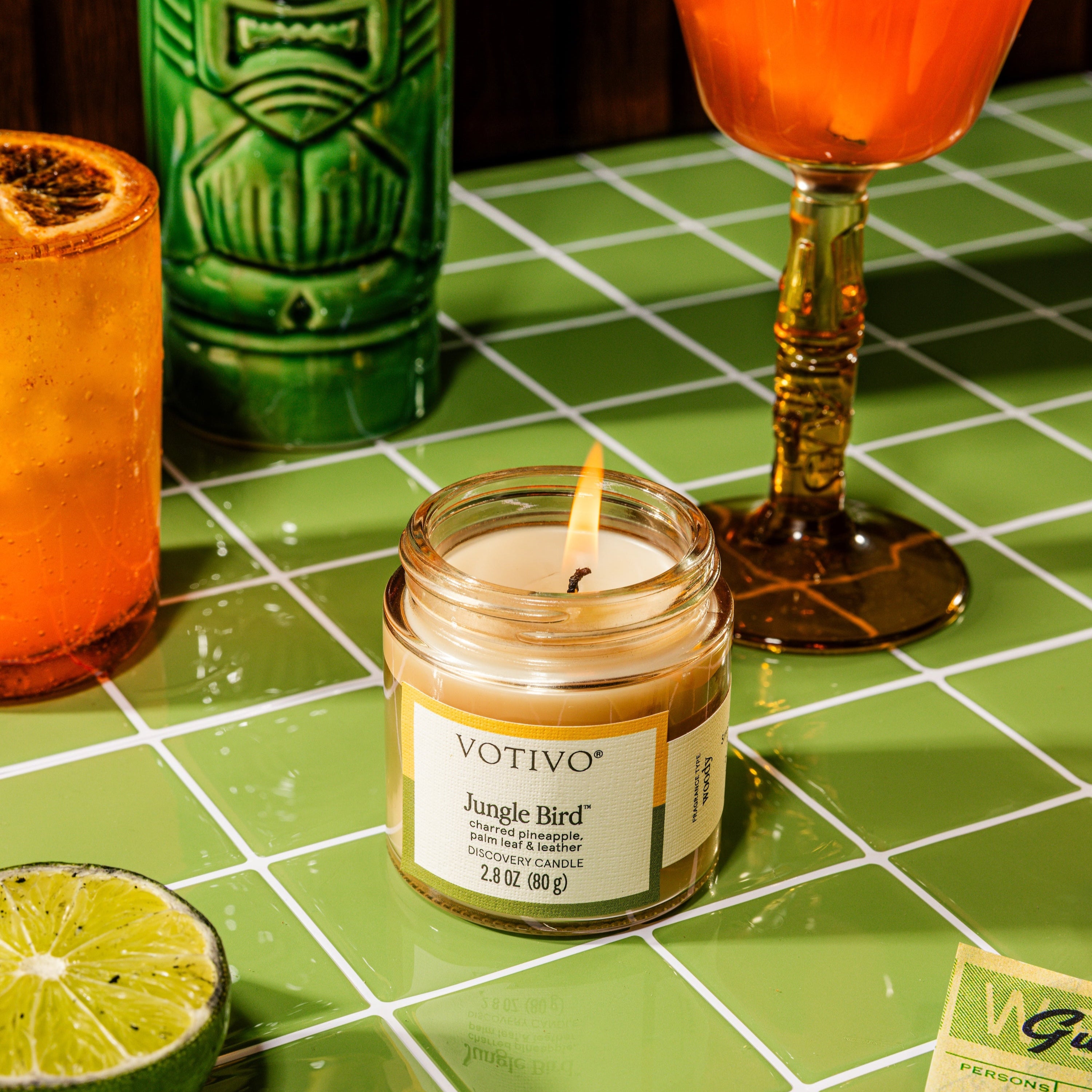 A lit Votivo Jungle Bird Discovery Candle sits on green tiles, surrounded by a lime wedge, tiki glass, orange cocktail, and sliced citrus. Warm light highlights its long-lasting fragrance.