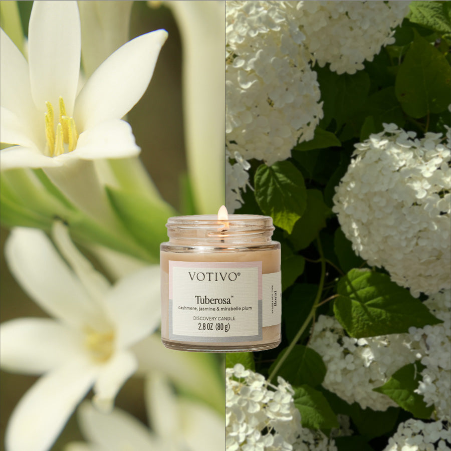 A lit Votivo Tuberosa Discovery Candle sits between white tuberose flowers and white hydrangea with green leaves, visually reflecting its signature tuberose fragrance.