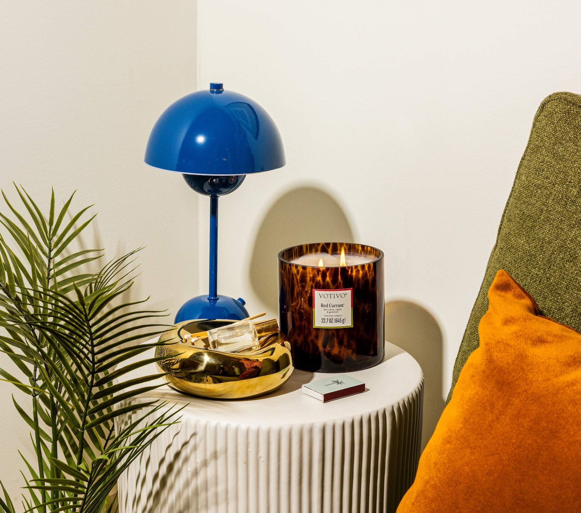 A blue lamp, Votivos Red Currant Tortoise Candle, a gold tray with matches, and a green sofa with an orange cushion rest on a white table by a small plant against a white wall.