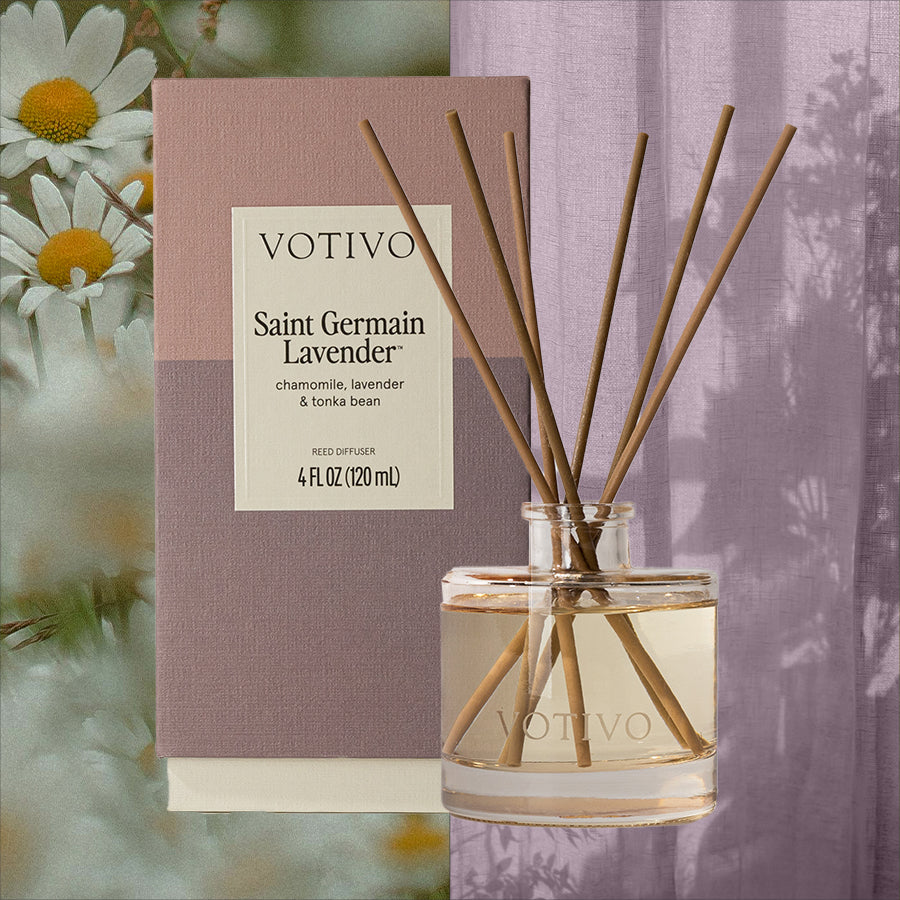 A Votivo Saint Germain Lavender Reed Diffuser with eight reeds sits in front of its lavender-toned box, while daisies and soft purple curtains in the background highlight its calming floral aroma and relaxing vibe.
