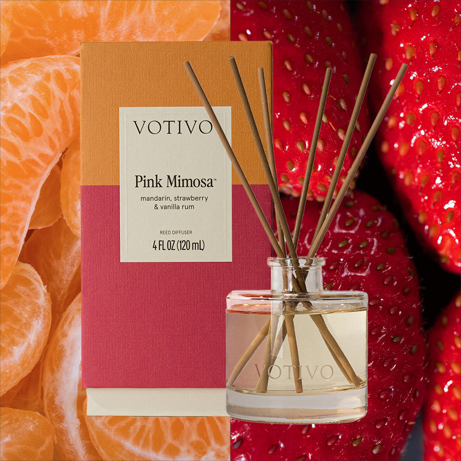 The Votivo Pink Mimosa Reed Diffuser comes with sticks in a glass bottle, set before its box. The backdrop features close-ups of mandarin oranges and strawberries, emphasizing the diffusers fruity scent.