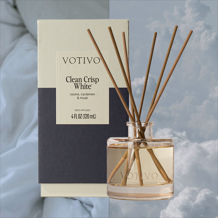 A Votivo Clean Crisp White Reed Diffuser with fiber reeds stands before its box, offering a continuous fragrance. The backdrop displays a cloudy sky.