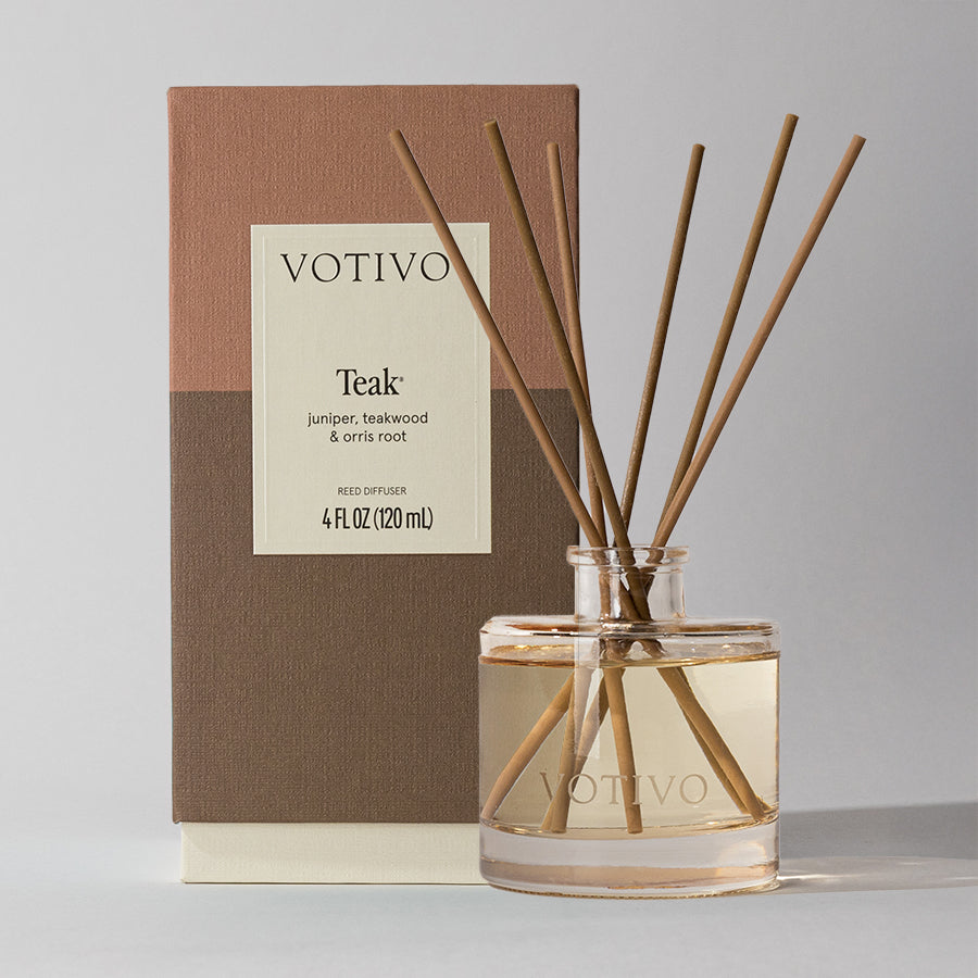 A Votivo Teak Reed Diffuser with fiber reeds and light brown liquid is shown in a clear glass bottle before its two-toned box. The label features a woody scent with notes of juniper, teakwood, and orris root.