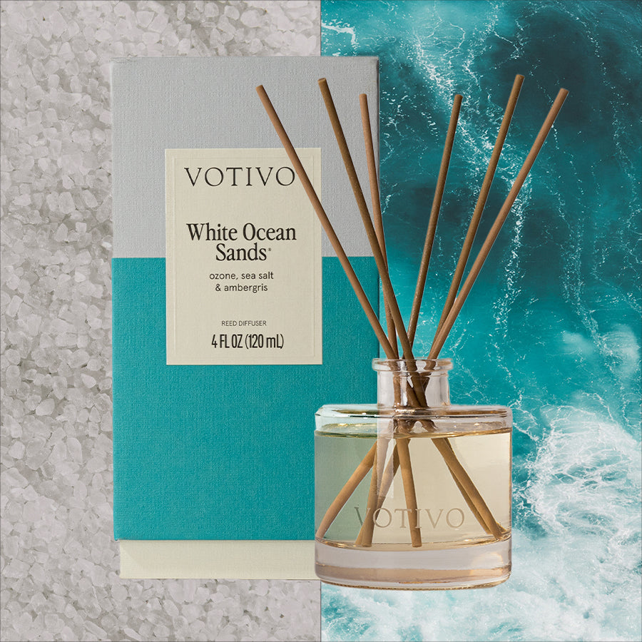 The Votivo White Ocean Sands Reed Diffuser, featuring brown reeds, is displayed with its blue-gray box against salt crystals and ocean waves.