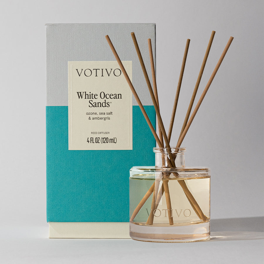 A clear Votivo glass jar with reeds is in front of a teal and gray White Ocean Sands Reed Diffuser box. The box lists notes of ozone, sea salt, and ambergris.