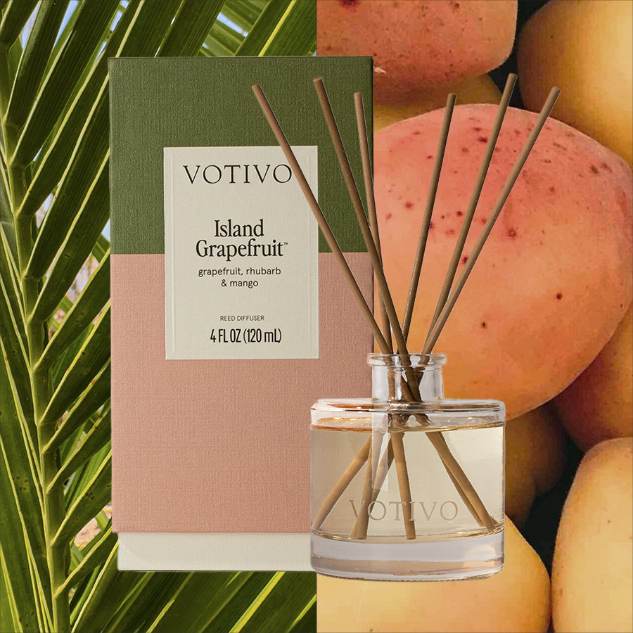 The Votivo Island Grapefruit Reed Diffuser and box are displayed with palm leaves and mangoes; the packaging highlights notes of grapefruit, rhubarb, and mango for a refreshing citrus fragrance.