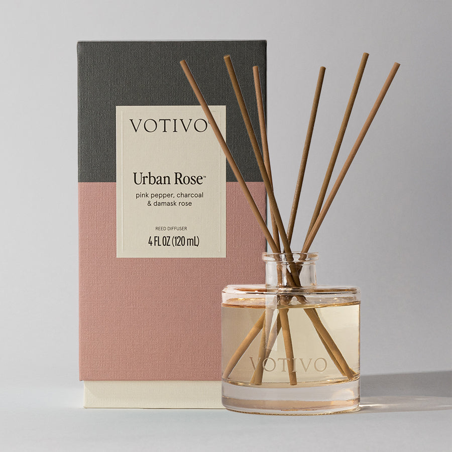 The Votivo Urban Rose Reed Diffuser, with eight fiber reeds in a clear glass bottle, is shown in front of its pink, grey, and cream box and features the Urban Rose scent: pink pepper, charcoal, and damask rose.