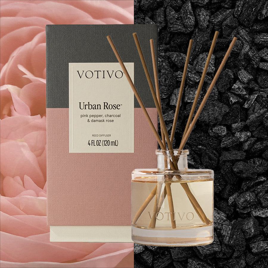 A Votivo Urban Rose Reed Diffuser with fiber reeds is displayed in front of its box, surrounded by pink rose petals and black charcoal. The packaging showcases the Urban Rose fragrance name and scent notes.