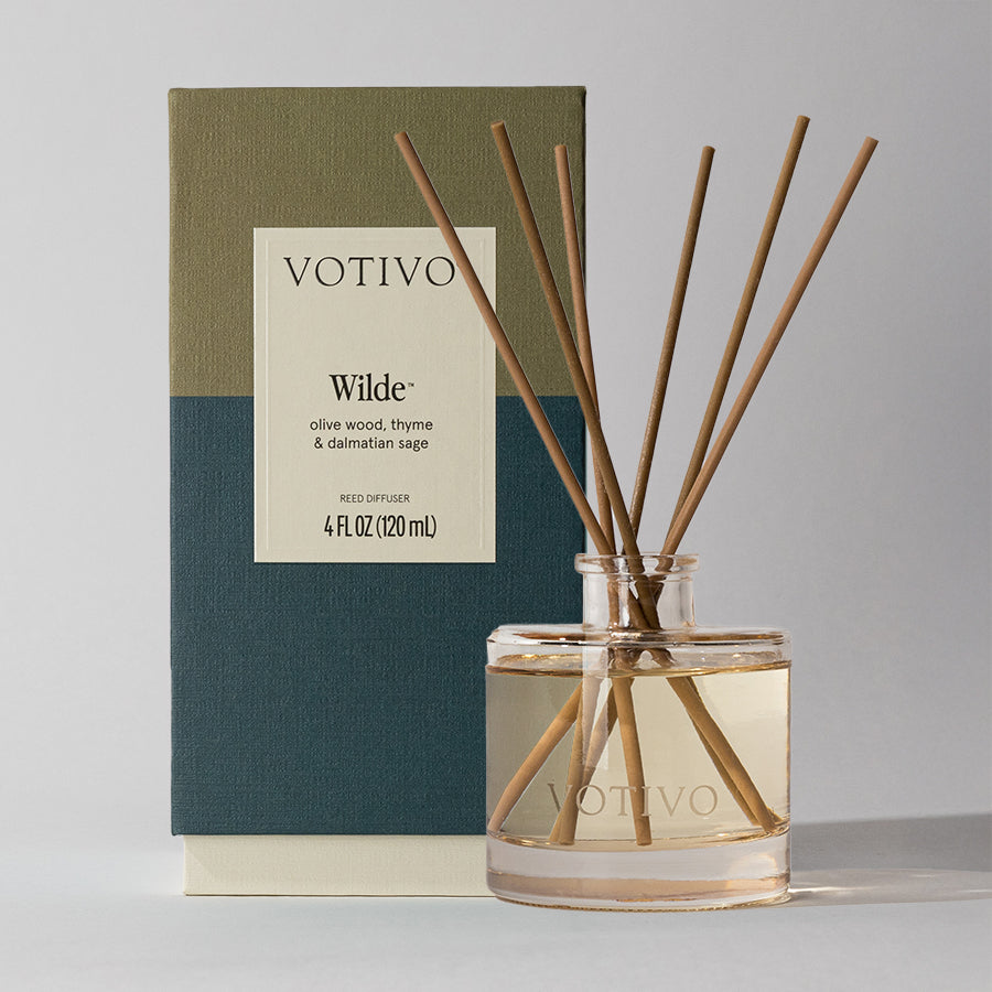 The Votivo Wilde Reed Diffuser features fiber reeds in a clear glass bottle for continuous fragrance and comes in a green and navy box labeled with “Votivo Wilde, olive wood, thyme & dalmatian sage.”.