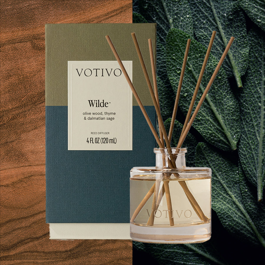 A Votivo Wilde Reed Diffuser with fiber reeds stands before its box. Wood grain and dark green sage leaves form the backdrop, while the label highlights olive wood, thyme, and dalmatian sage for a continuous fragrance experience.