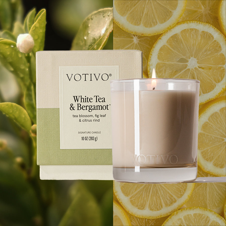 A Votivo White Tea & Bergamot 10 oz. Signature Candle is displayed beside its box, accented by fresh green leaves and lemon slices to showcase its citrus and herbal fragrance. Made with a clean Soy Blend Wax for quality burning.
