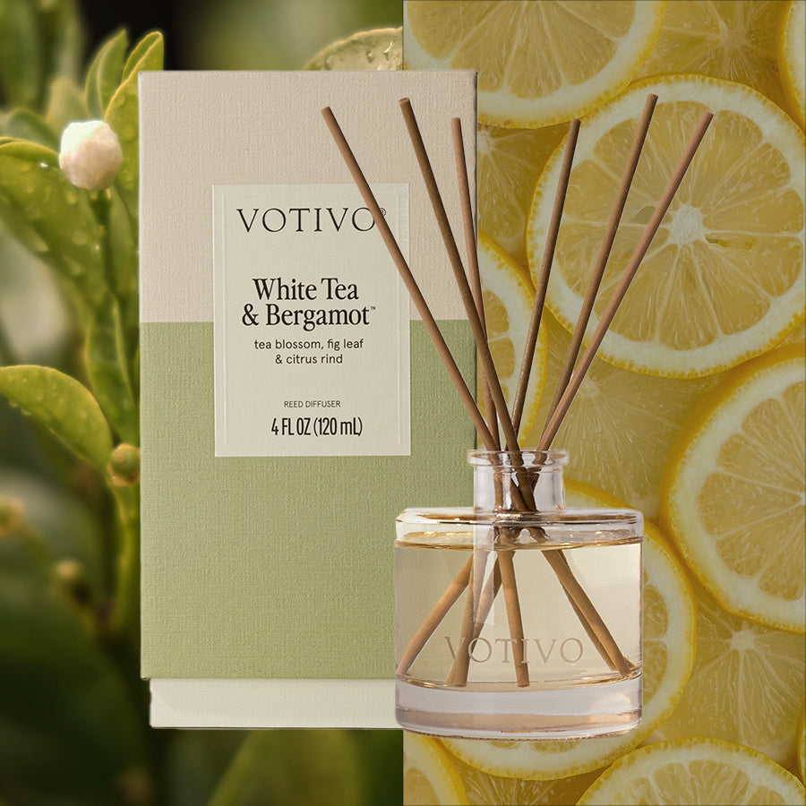 A Votivo White Tea + Bergamot Reed Diffuser is displayed with its pale green and cream packaging and several fiber reeds, placed in front of lemons and green foliage. The diffuser contains clear liquid.