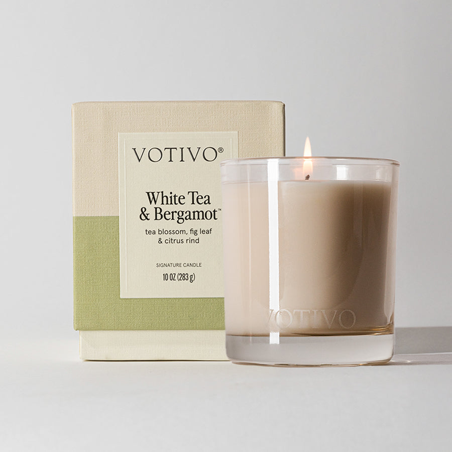 The Votivo White Tea & Bergamot 10 oz. Signature Candle features a premium soy blend for a clean, long-lasting burn and comes in a clear glass holder beside its beige and green box listing scent notes and size.