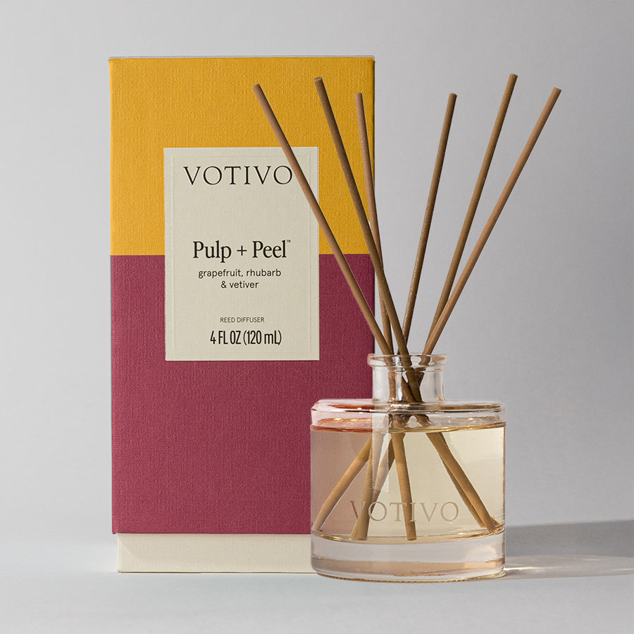 A Votivo Pulp + Peel Reed Diffuser with fiber reeds and pale liquid stands in front of its yellow and burgundy box labeled Pulp + Peel, releasing a continuous scent of grapefruit, rhubarb, and vetiver into the air.