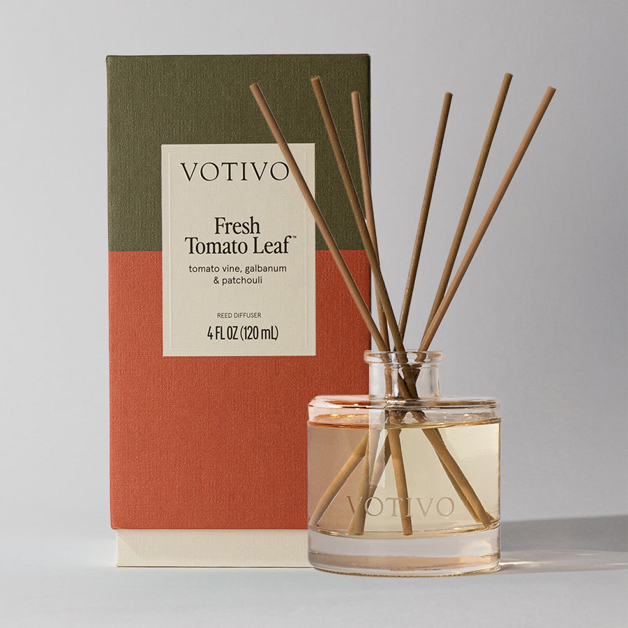 A Votivo Fresh Tomato Leaf Reed Diffuser with several fiber reeds stands before its green and orange box, the clear liquid visible inside the elegant bottle.