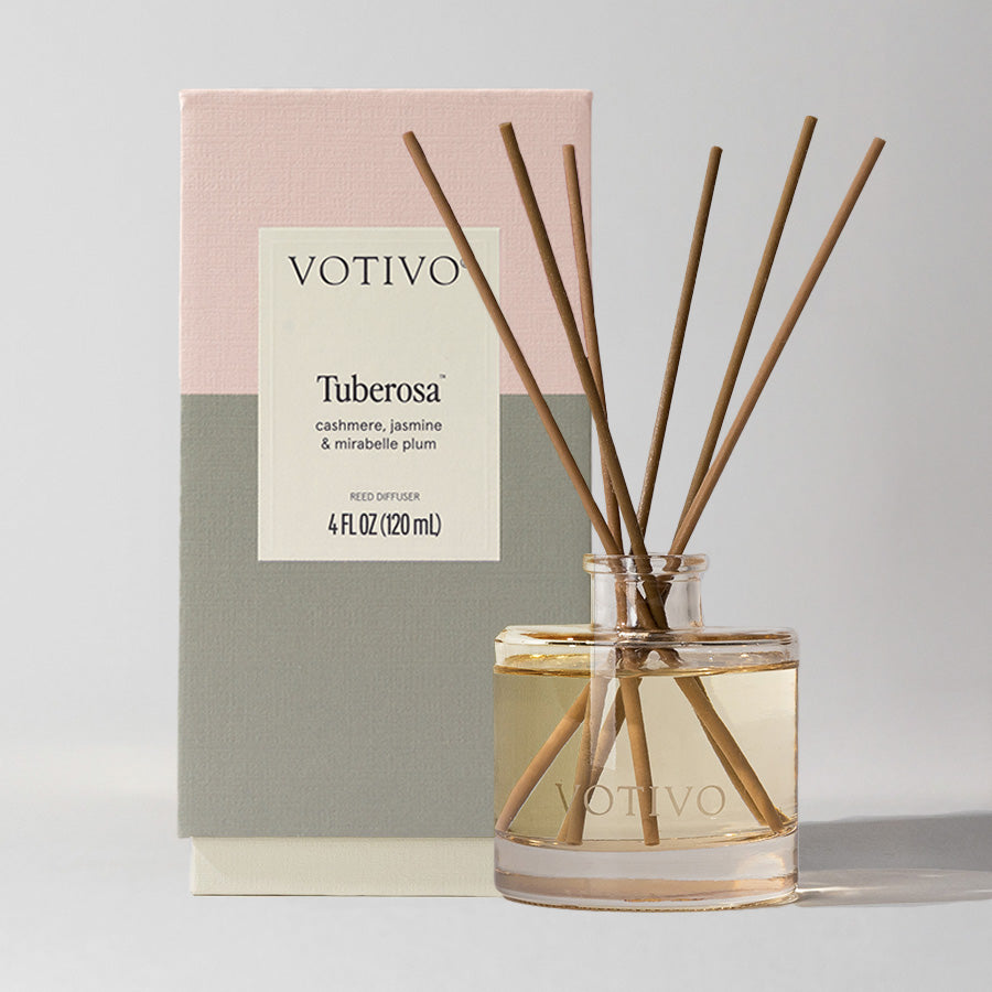 The Votivo Tuberosa Reed Diffuser features fiber reeds in a clear glass bottle with a Tuberose scent, displayed before a pink, cream, and gray box. The label highlights notes of cashmere, jasmine, and mirabelle plum.