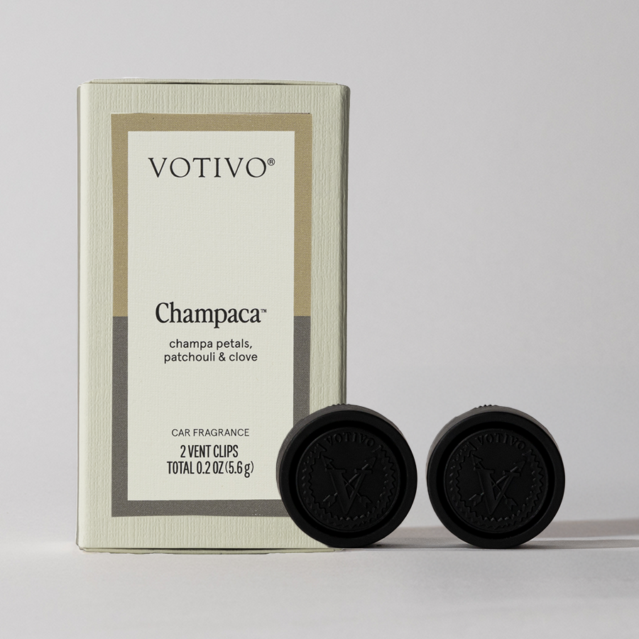 A Votivo Champaca Car Fragrance (2 Pack) box stands upright next to two black vent clips, highlighting a woody scent of champa petals, patchouli, and clove. Each box contains two vent clips for car fragrance.