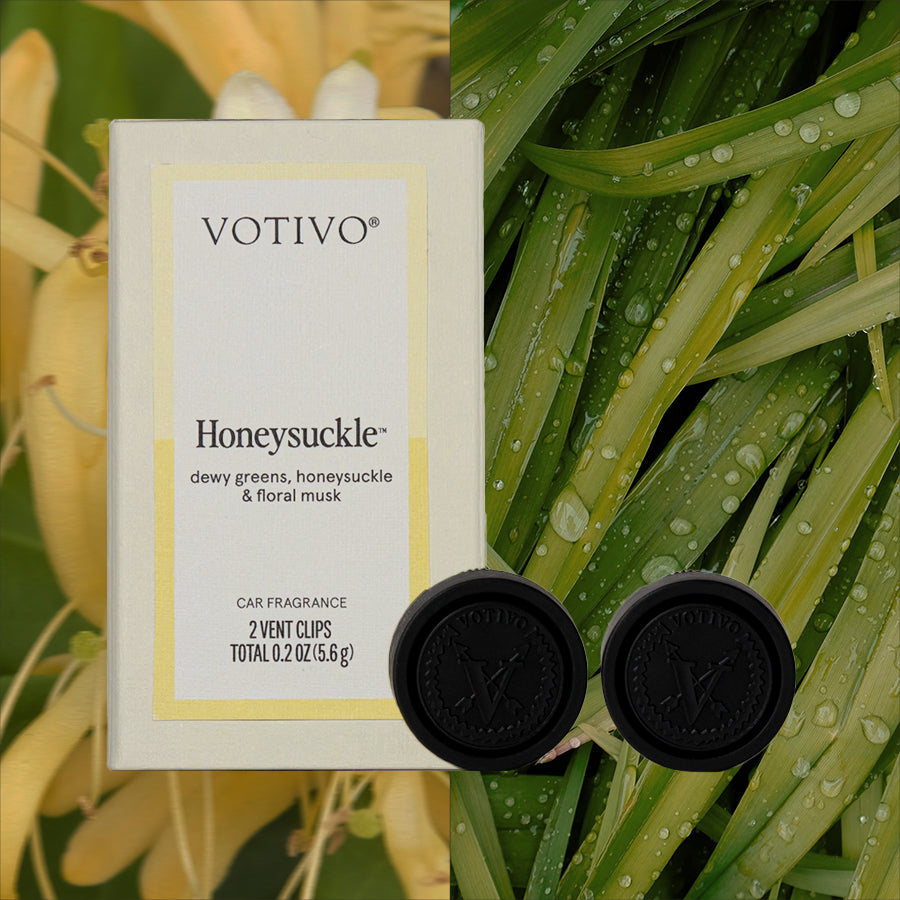 A box of Votivo Honeysuckle Car Fragrance (2 Pack) and two black vent clips are displayed against dew-kissed leaves and yellow honeysuckle flowers, evoking a fresh honeysuckle scent for your car.