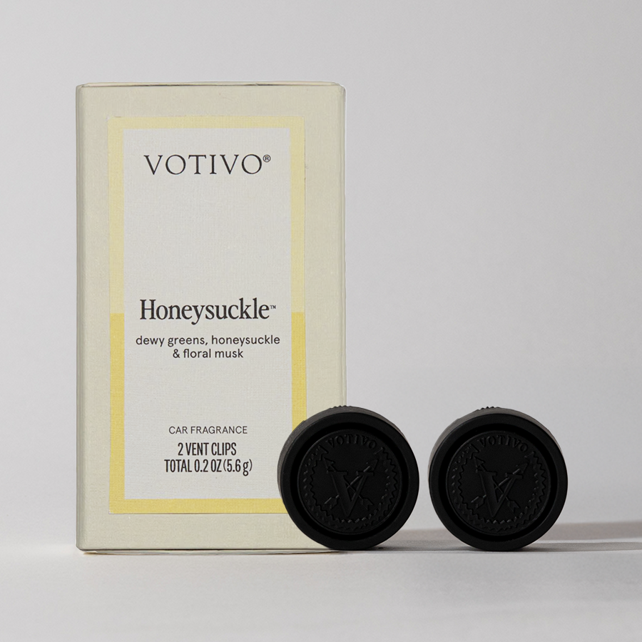 A cream-colored Votivo Honeysuckle Car Fragrance (2 Pack) box with gold accents stands upright beside two black vent clips, all set against a neutral background, hinting at the inviting floral scent inside.