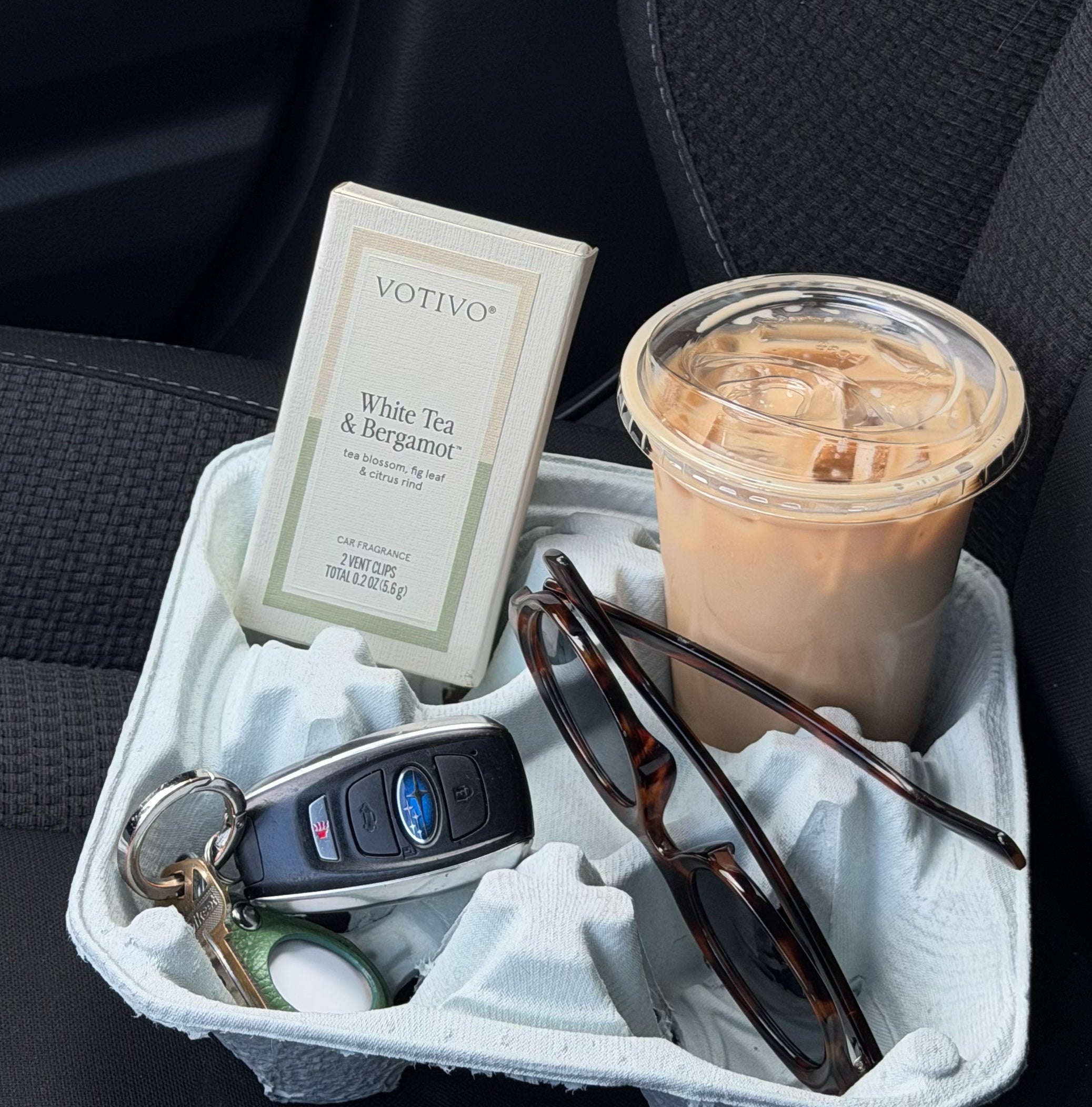 A key fob, sunglasses, iced coffee, and a box of Votivo White Tea & Bergamot Car Fragrance (2 Pack)—ideal for paraben free scent on-the-go—rest in a cup holder inside the car.