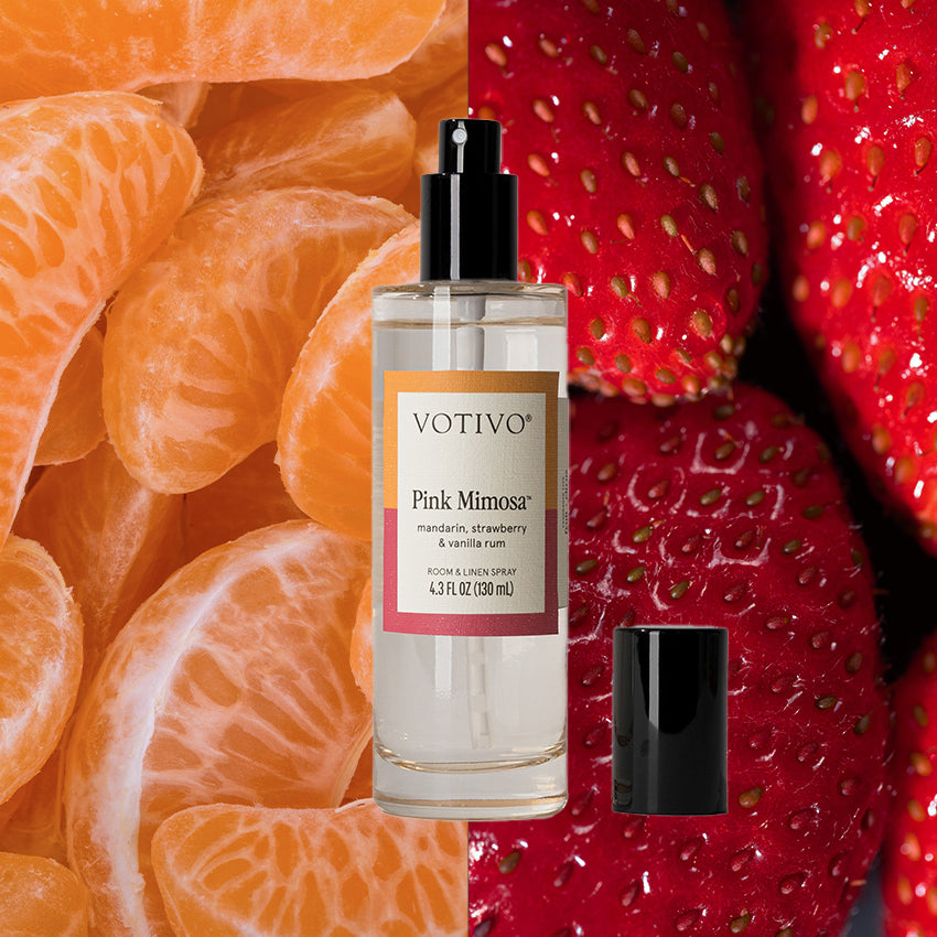 A bottle of Votivo Pink Mimosa Room & Linen Spray stands upright with its cap beside it, set against a backdrop of peeled mandarin orange segments on the left and ripe strawberries on the right, highlighting its fresh, fruity fragrance.