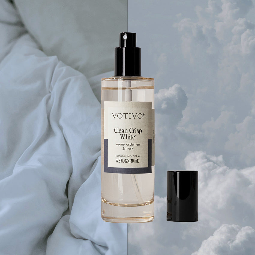 A Votivo Clean Crisp White Room & Linen Spray bottle with a black cap is displayed against a split background of light blue bed sheets and a cloudy sky.