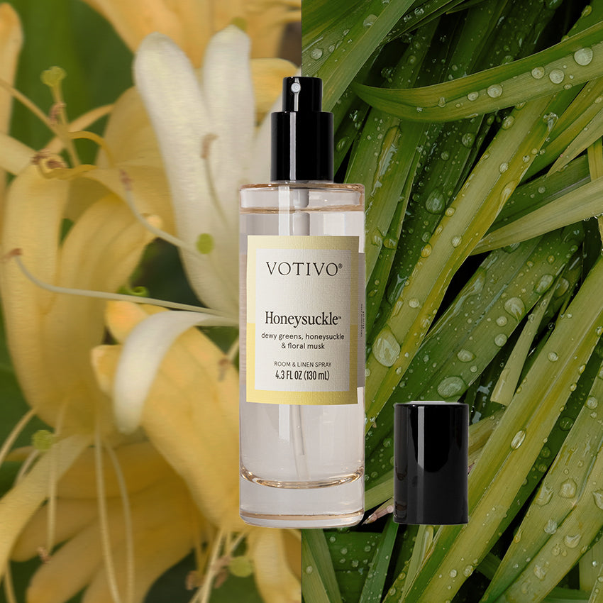A Votivo Honeysuckle Room & Linen Spray with a black cap is set against a backdrop of yellow honeysuckle flowers on the left and green leaves on the right, capturing its fresh floral scent.